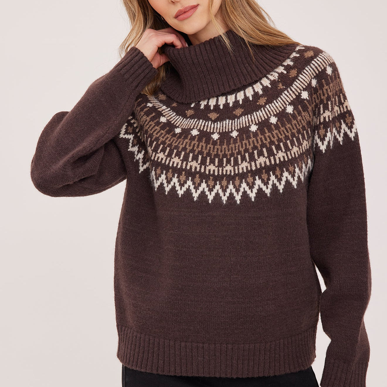 A woman wearing a dark brown knit sweater with a white and beige Fair Isle yoke pattern, paired with black pants, against a light gray studio background.