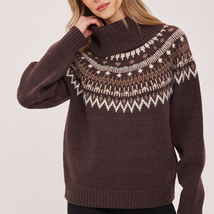 A woman wearing a dark brown knit sweater with a white and beige Fair Isle yoke pattern, paired with black pants, against a light gray studio background.